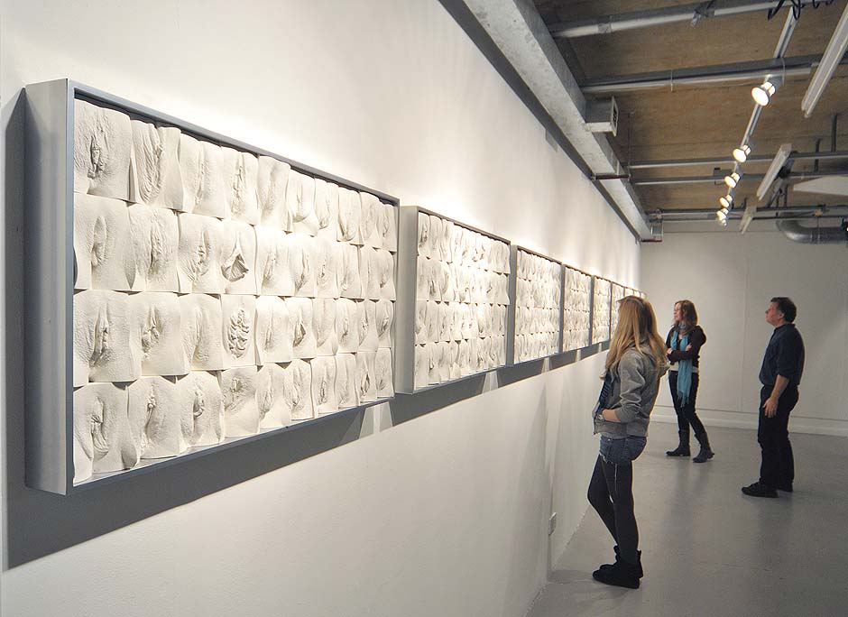 Jamie Mc Cartney, The great wall of vagina, exhibition view with visitors. The 9 metre long polyptych consists of four hundred plaster casts of vulvas, all of them unique, arranged into ten large panels.